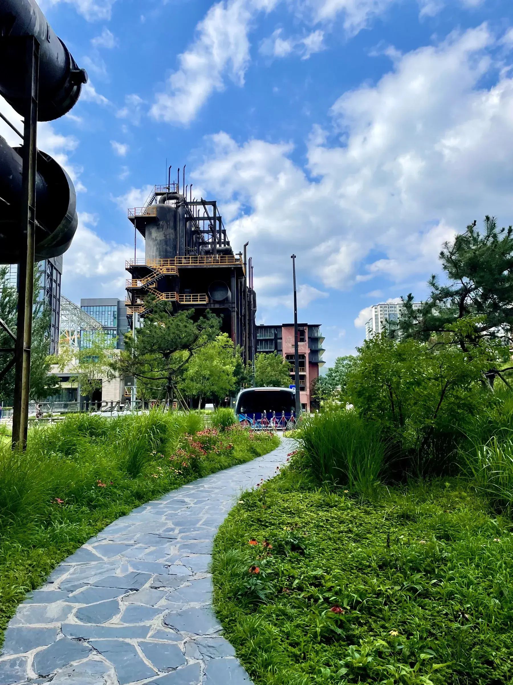 Stone garden path lined with flowers leading toward a dark industrial tower at Shougang Park, Beijing