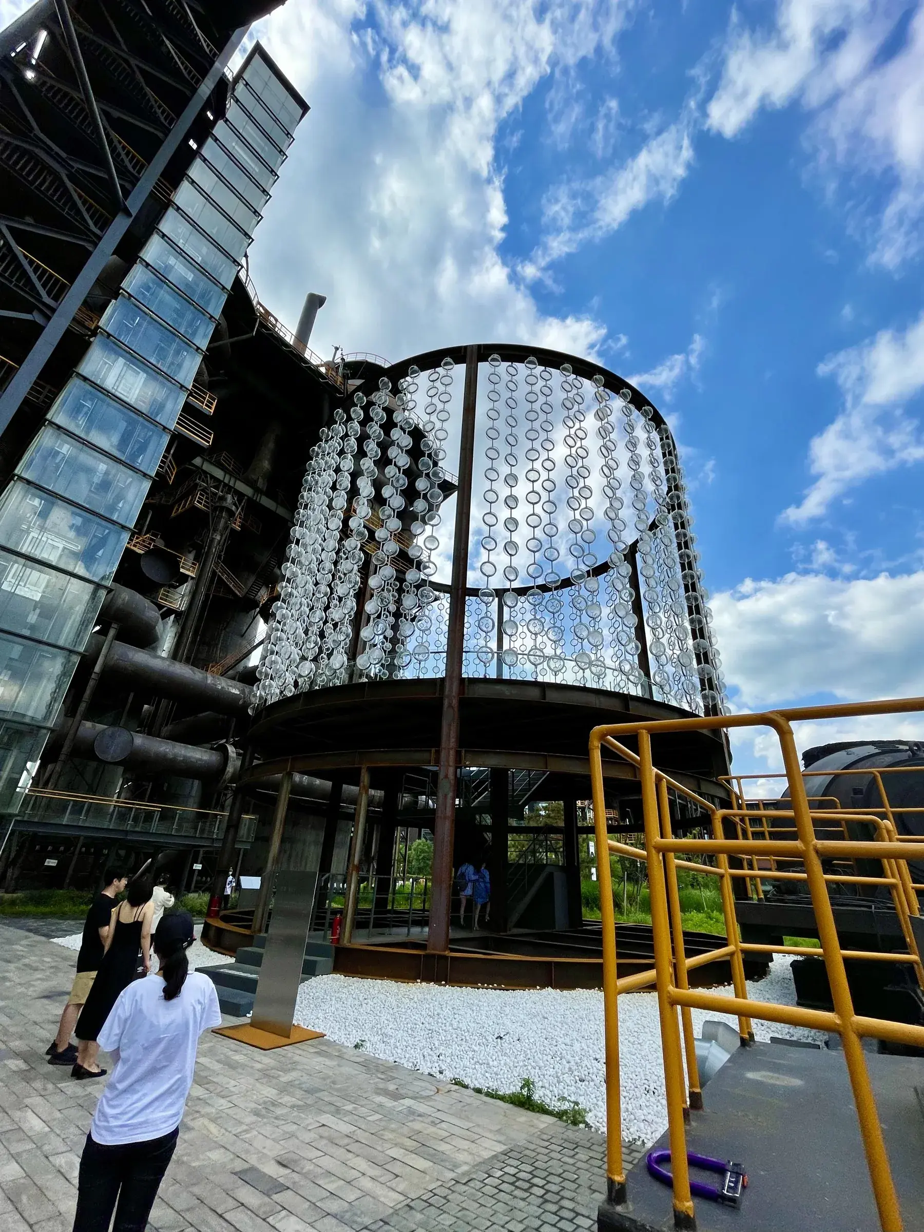 Rusted cylindrical steel structure hung with translucent glass discs beside a yellow staircase at Shougang Park, Beijing