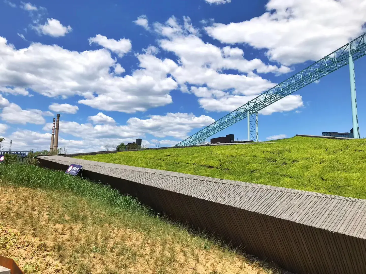 Grassy embankment and ribbed stone wall beneath the light-blue steel gantry at Shougang Park, Beijing