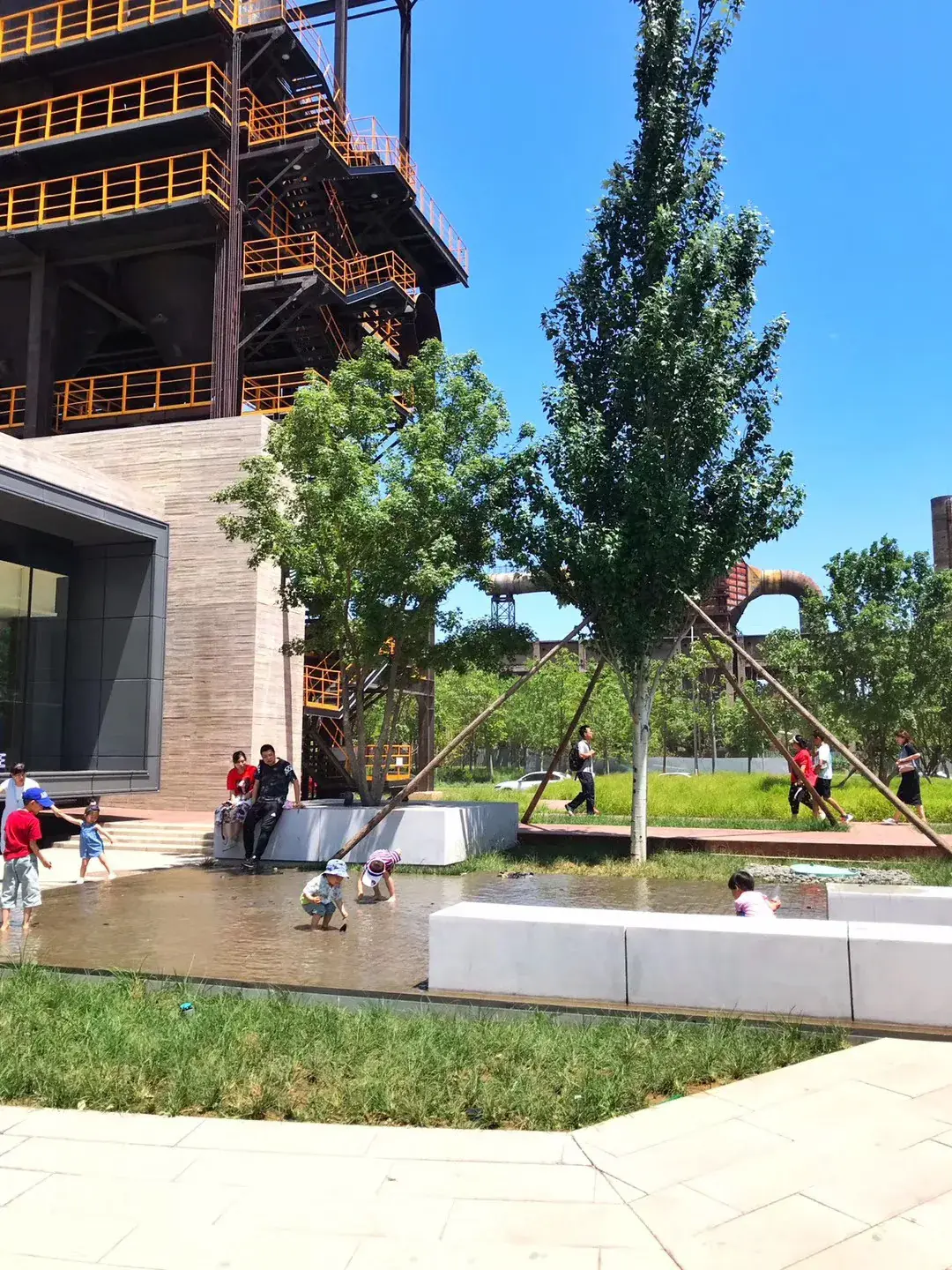 Children playing in a shallow plaza water feature beneath orange-railed industrial walkways at Shougang Park, Beijing