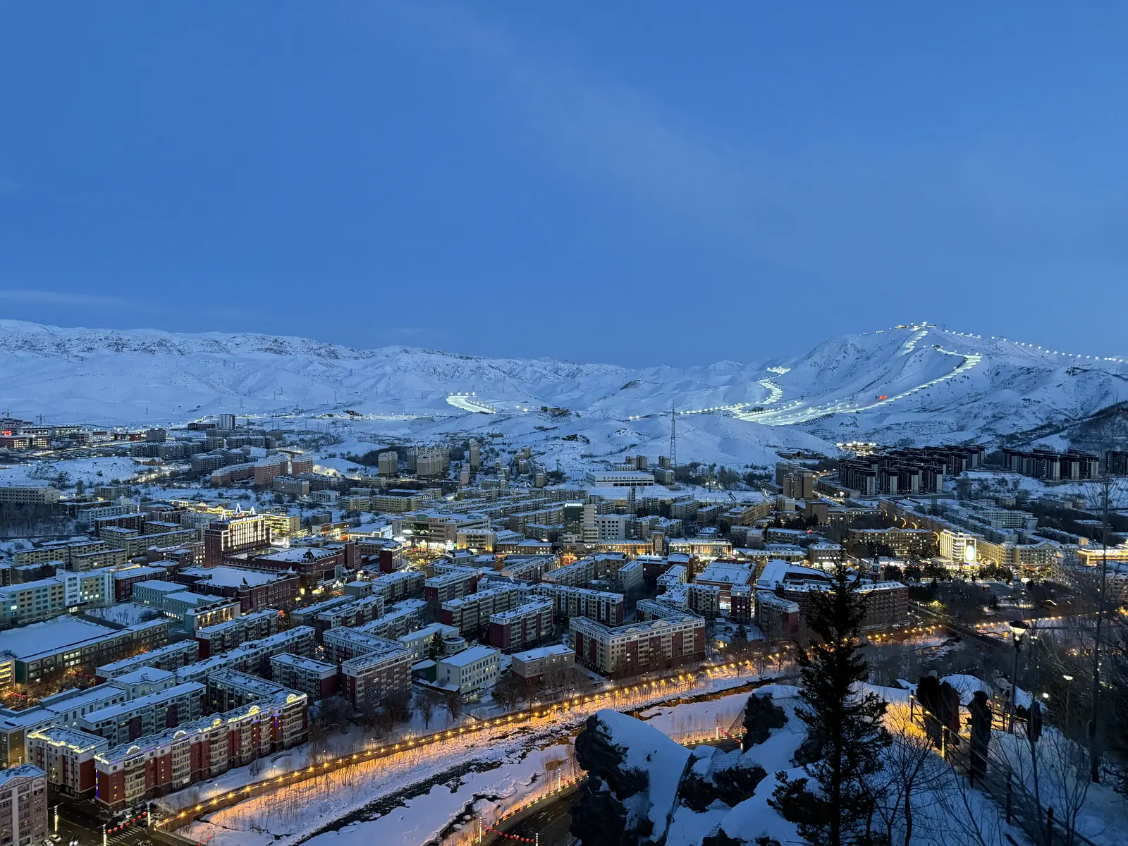 Floodlit Jiangjunshan ski runs above snow-covered Altay at blue hour in December 2025.