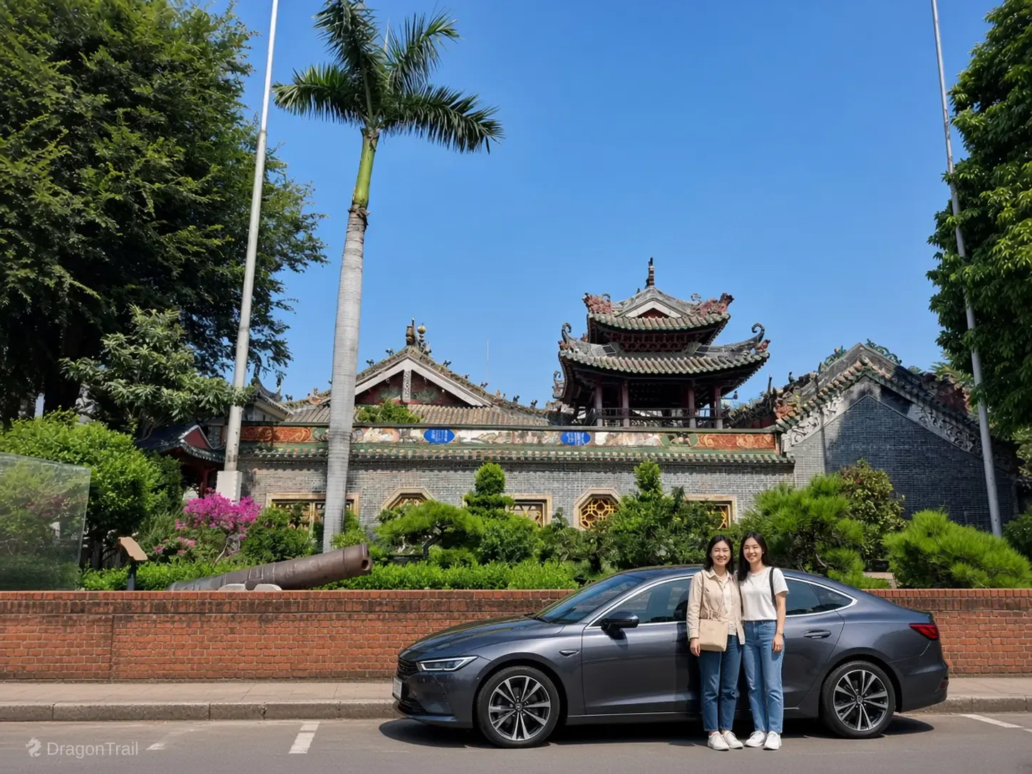 Two women stand beside a dark sedan on the street outside the Foshan Ancestral Temple, with the tiled roofs and garden wall behind them